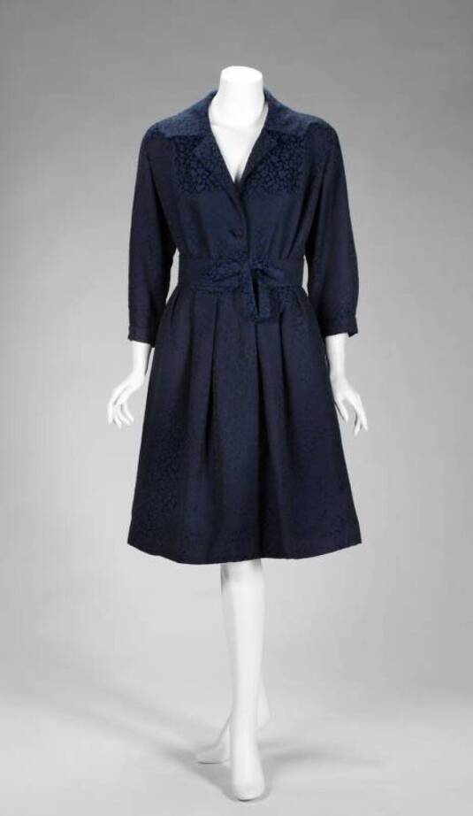GRETA GARBO navy blue and black dress: An early 1950s dress by Valentina in navy blue satin and wool jacquard with a black abstract pattern. The dress has a V-neck with notched collar, single button closure on front of dress and zipper at back. The skirt has some box pleating. Accompanied by a structured matching belt embellished with a bow. 